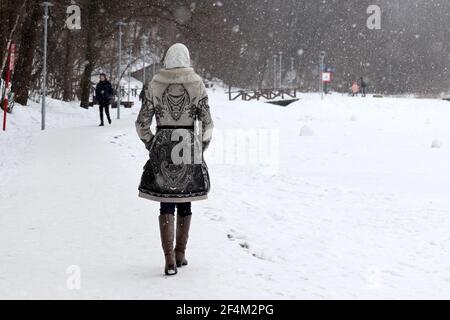 Lonely ragazza in pelliccia cappotto che cammina nel parco invernale durante la nevicata. Clima freddo in primavera, vista pittoresca sugli alberi ricoperti di neve e sul lago ghiacciato Foto Stock