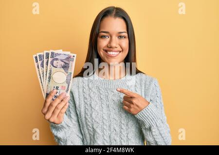 Bella donna ispanica che tiene 5000 yen giapponesi banconote sorridente felice puntare con la mano e il dito Foto Stock