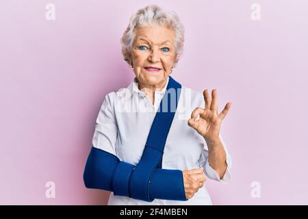 Donna senior con capelli grigi che indossa il braccio sull'imbragatura facendo ok segno con le dita, sorridente amichevole gesturing simbolo eccellente Foto Stock