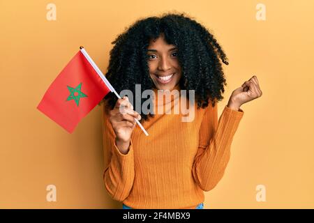 Donna afroamericana con i capelli afro che tengono bandiera del marocco urlando orgoglioso, celebrando la vittoria e il successo molto eccitato con il braccio sollevato Foto Stock