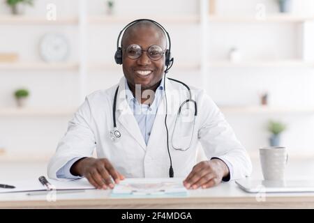 Concetto di sanità e medicina. Allegro medico afroamericano sorridente alla macchina fotografica, seduto al suo posto di lavoro in ufficio Foto Stock