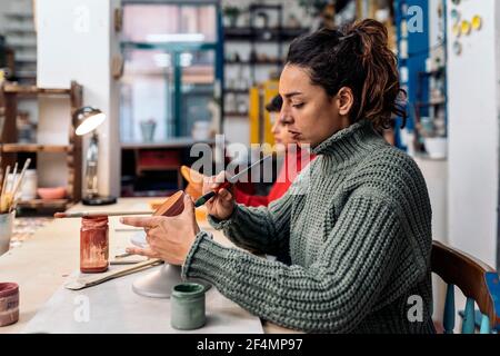 Foto di stock di donna messa a fuoco utilizzando pennello durante la sua classe di ceramica. Foto Stock