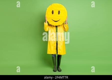 Foto a corpo pieno di giovane uomo vicino copertina carta pinata emoji smile indossare impermeabile isolato su sfondo di colore verde Foto Stock