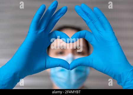Primo piano di un'infermiera irriconoscibile con maschera medica e guanti protettivi. Sta formando un cuore con le mani. Concetto di salute. Messa a fuoco selettiva. Foto Stock