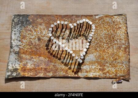 Cuore di chiodi martellato in stagno arrugginito appiattito su tavola di legno. Foto Stock