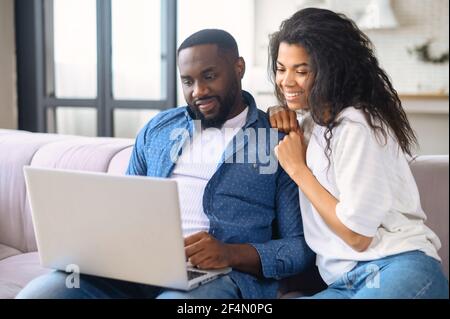 Una simpatica coppia afro-americana trascorre il tempo libero online con un computer portatile a casa, una donna affascinante e un bell'uomo seduto sul divano, guardando film o facendo acquisti online insieme Foto Stock