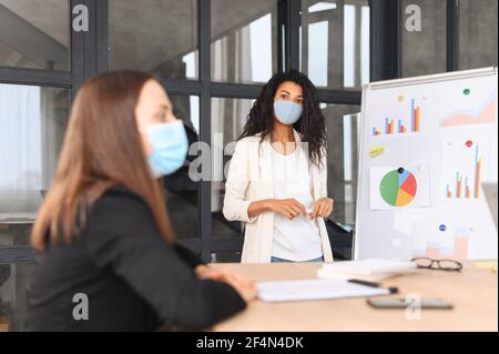 Gruppo di lavoro multietnico brainstorming in ufficio contemporaneo durante il periodo pandemico, un diversi colleghi indossare maschera medica al coperto. Una donna afroamericana con faccia coperta vicino a lavagna bianca Foto Stock