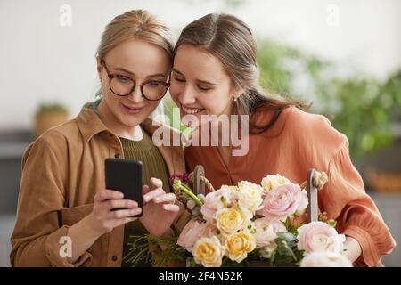 Ritratto in vita di due giovani donne che guardano lo smartphone schermo e sorridente mentre si lavora in negozio di fiori Foto Stock