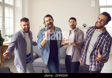 Sorridendo eccitato giovani amici uomini cantare canzoni in microfono e. avere festa a casa Foto Stock