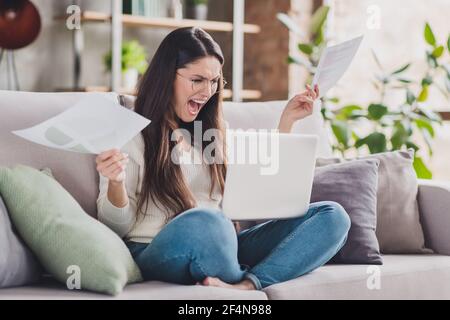 Foto della signora pazza stressata tenere carte computer urlare aperto bocca indossare occhiali bianco pullover in soggiorno casa al chiuso Foto Stock