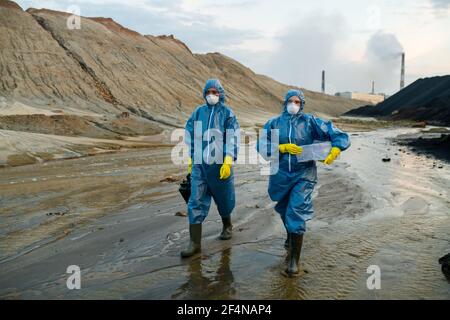 Due giovani scienziati contemporanei in tute protettive blu e. stivali in gomma che si muovono lungo il fiume inquinato circondato da colline Foto Stock