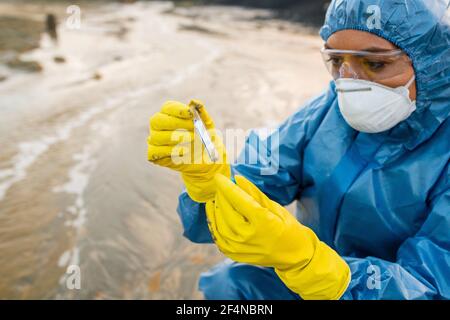 Giovane ecologa femminile in respiratore e occhiali guardando la fiasca con campione di acqua sporca o suolo in inquinato e. area industriale tossica Foto Stock
