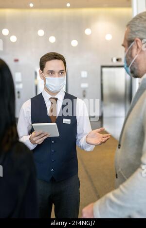 Giovane amministratore maschile dell'hotel in maschera protettiva e abbigliamento da formalwear utilizzo del touchpad durante la conversazione con nuovi ospiti nella sala Foto Stock