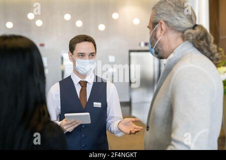 Giovane receptionist maschile in maschera protettiva e abbigliamento da formalwear con touchpad mentre si parla con un uomo d'affari maturo nella sala Foto Stock