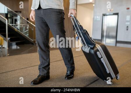 Sezione bassa del moderno uomo d'affari in casualwear tirando la valigia dietro si sposta lungo la hall di un grande e lussuoso hotel Foto Stock