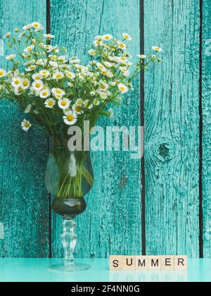 Beautiful bouquet of forest daisy in vintage glass vase opposite old turquoise wooden wall. Word SUMMER from wooden letters Foto Stock