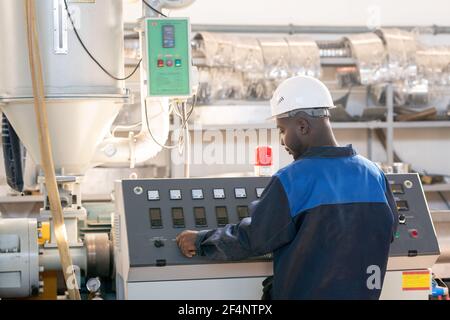 Vista posteriore di giovane operaio africano maschile in tute e. hardhat si trova di fronte al pannello di controllo di enormi industrie macchina e manopola di rotazione Foto Stock