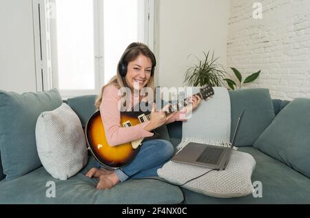 Attraente musicista donna che suona la chitarra elettrica durante il concerto online a casa in isolamento. Insegnante di musica che insegna strumento di gioco online usando l Foto Stock
