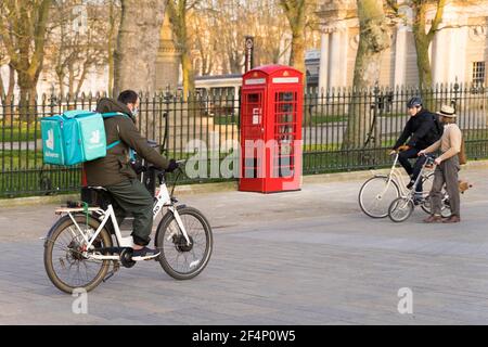 Deliveroo uomo sulla sua bici del corriere sul relativo senso al cliente di takeaway, scatola rossa del telefono e due altri ciclisti, Londra Greenwich Inghilterra Foto Stock
