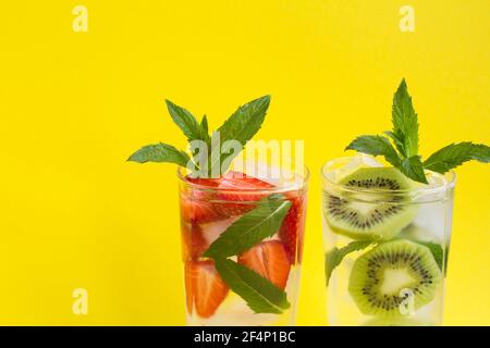 Infondere acqua o limonata con fragola, kiwi, ghiaccio e menta nei due bicchieri su sfondo giallo. Primo piano. Spazio di copia. Foto Stock