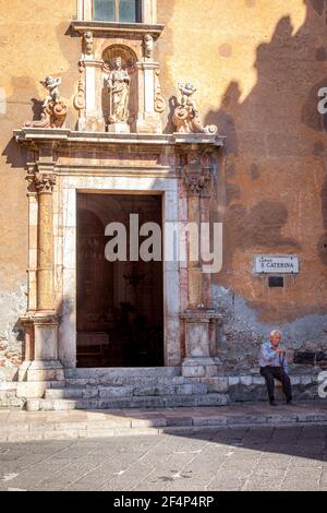 Uomo anziano seduto alla porta di fronte alla Chiesa di Santa Caterina (b. 1610) a Taormina, Sicilia, Italia Foto Stock