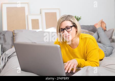 Donna sorridente in pullover giallo e jeans è sdraiato sul letto a casa con il laptop, lavorando online, guardando le notizie. Concetto di tecnologia. Foto Stock