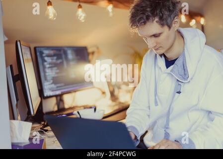 Un uomo seduto su una scrivania che lavora da casa sul suo computer. Programmatore SPECIALIZZATO IT che scrive il codice sul suo pc e laptop. Ufficio domestico remoto dello sviluppatore Foto Stock