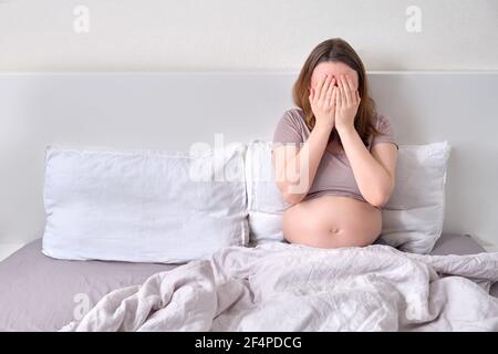 Una donna incinta solitaria si trova nel letto coprendo il viso con le mani. Problemi di gravidanza e solitudine Foto Stock