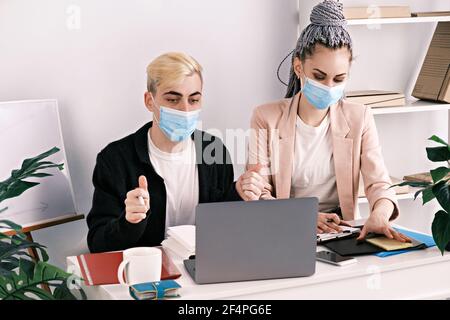 Conferenza business online. Giovane manager maschile e il suo assistente in giubbotti e maschere mediche utilizzando un computer portatile per tenere una conferenza. Segnalazione ai dirigenti Foto Stock