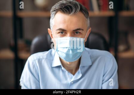 Ritratto di un uomo caucasico di mezza età dai capelli grigi, lavoratore d'ufficio, manager, business leader in una camicia elegante, indossando una maschera medica di protezione, guardando la fotocamera. Concetto di assistenza sanitaria Foto Stock