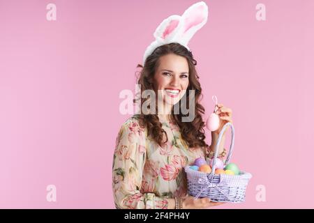 donna trendy di 40 anni in abito floreale con orecchie conigliate e cesto di uova di pasqua isolato su sfondo rosa. Foto Stock