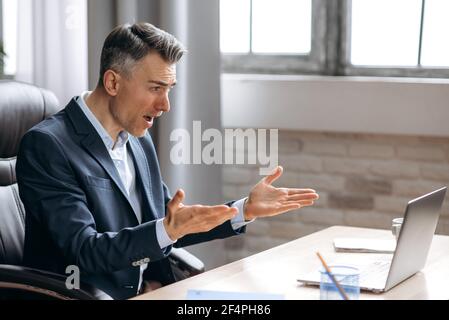 Scioccato infastidito caucasico impegnato uomo d'affari in tuta, guardando un portatile in confusione, ricevuto notizie inaspettate, fallito un accordo, grida, gesturing con le mani, seduto alla scrivania dell'ufficio Foto Stock