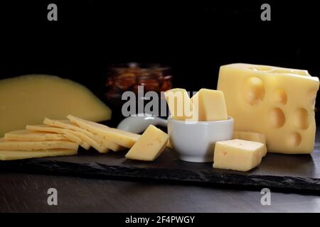 Diversi tipi di formaggio in pezzi, tagliati a fette e pezzi in ciotola bianca con cucchiaio e vaso di noci sono disposti su tavola di pietra nera sul tavolo. Che Foto Stock