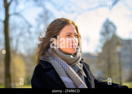 Attraente donna di mezza età che indossa una sciarpa calda e un cappotto rilassante all'aperto sotto il sole primaverile seduto su una panca del parco dentro un primo piano testa e spalla Foto Stock