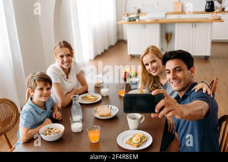 Gruppo di quattro persone caucasiche che scatta foto selfie mentre si mangia una colazione sul tavolo da pranzo. Padre, Madre, figlia e Figlio godendo del tempo felice insieme con la cucina in background. Foto Stock