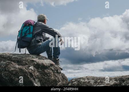 Turista con zaino siede sulla cima della montagna, riposando e ammirando la vista dall'alto. Il concetto di attività all'aperto, viaggio e avventura. Foto Stock