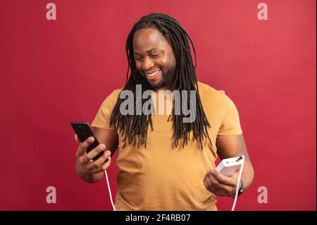 Felice ragazzo afroamericano soddisfatto con i Dreadlock in piedi isolato sopra la parete rossa e tenendo il telefono cellulare collegato al power bank tramite cavo USB, in carica, utilizzando il cellulare cellulare, sorridente Foto Stock