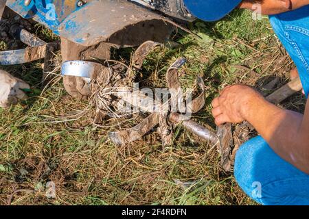 Un agricoltore irriconoscibile ripara un aratro motore portatile. Macchine agricole: Coltivatore per dissodamento in giardino, aratro a mano motorizzato. Autentico Foto Stock