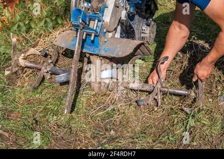 Un agricoltore irriconoscibile ripara un aratro motore portatile. Macchine agricole: Coltivatore per dissodamento in giardino, aratro a mano motorizzato. Autentico Foto Stock