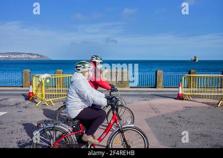 Due ciclisti donne che usano biciclette elettriche che cavalcano il prom di Douglas, l'Isola di Man che indossa maschere in Covid Times Foto Stock