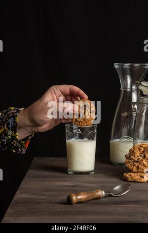 primo piano della mano della donna con biscotti al cioccolato in mano su un bicchiere di latte, su un tavolo con bottiglia di latte e biscotti, fondo nero, verticale Foto Stock