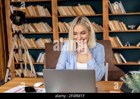 Bella donna di affari sta avendo una videoconferenza con i colleghi, sorridente. Donna di successo con capelli biondi seduta alla scrivania in ufficio a casa Foto Stock