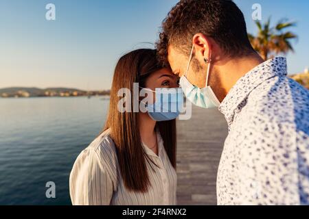 L'uomo e la donna guardano gli occhi l'uno contro l'altro appoggiando le fronte l'uno contro l'altro indossando una maschera protettiva a causa di Coronavirus. Giovane coppia di turisti Foto Stock