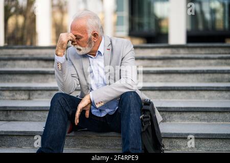 L'uomo d'affari anziano è stanco di lavoro. È seduto di fronte alla costruzione dell'azienda e ha mal di testa. Foto Stock