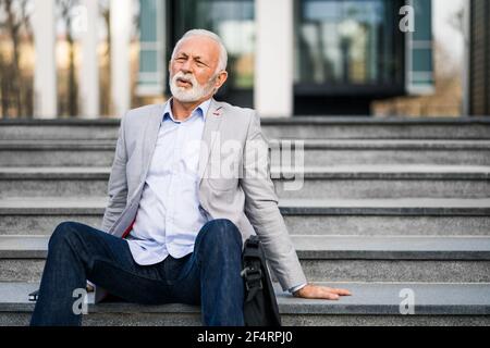 L'uomo d'affari anziano è stanco di lavoro. È seduto sulle scale di fronte all'edificio aziendale. Foto Stock