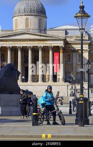 Londra, Inghilterra, Regno Unito. Trafalgar Square. Deliveroo delivery man su una bici elettrica EFF. Galleria Nazionale dietro Foto Stock