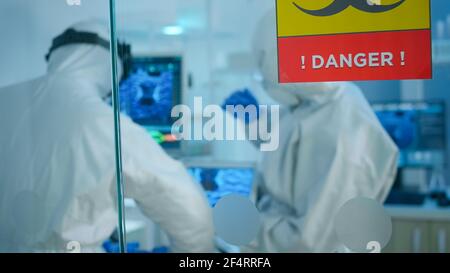 Chimici in tuta cercando di sviluppare il vaccino utilizzando la compressa in piedi dietro la parete di vetro che lavora nella zona di pericolo del laboratorio. Gli scienziati che esaminano l'evoluzione del virus ricercando il trattamento contro il covid19 Foto Stock