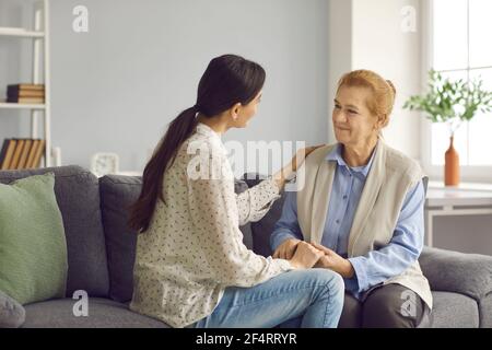 Felice madre anziana e giovane figlia seduta sul divano, tenendo le mani e parlando Foto Stock