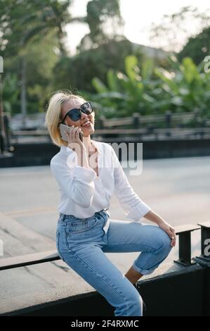 Una giovane donna asiatica seduta in strada sorride felice mentre parla sul suo cellulare. Buona sensazione di spensieratezza. Foto Stock