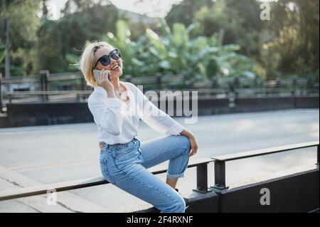 Una giovane donna asiatica seduta in strada sorride felice mentre parla sul suo cellulare. Buona sensazione di spensieratezza. Foto Stock